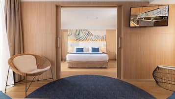 A modern hotel room with a large bed, a wicker chair, a flat-screen TV, and a view of the cityscape through the window.