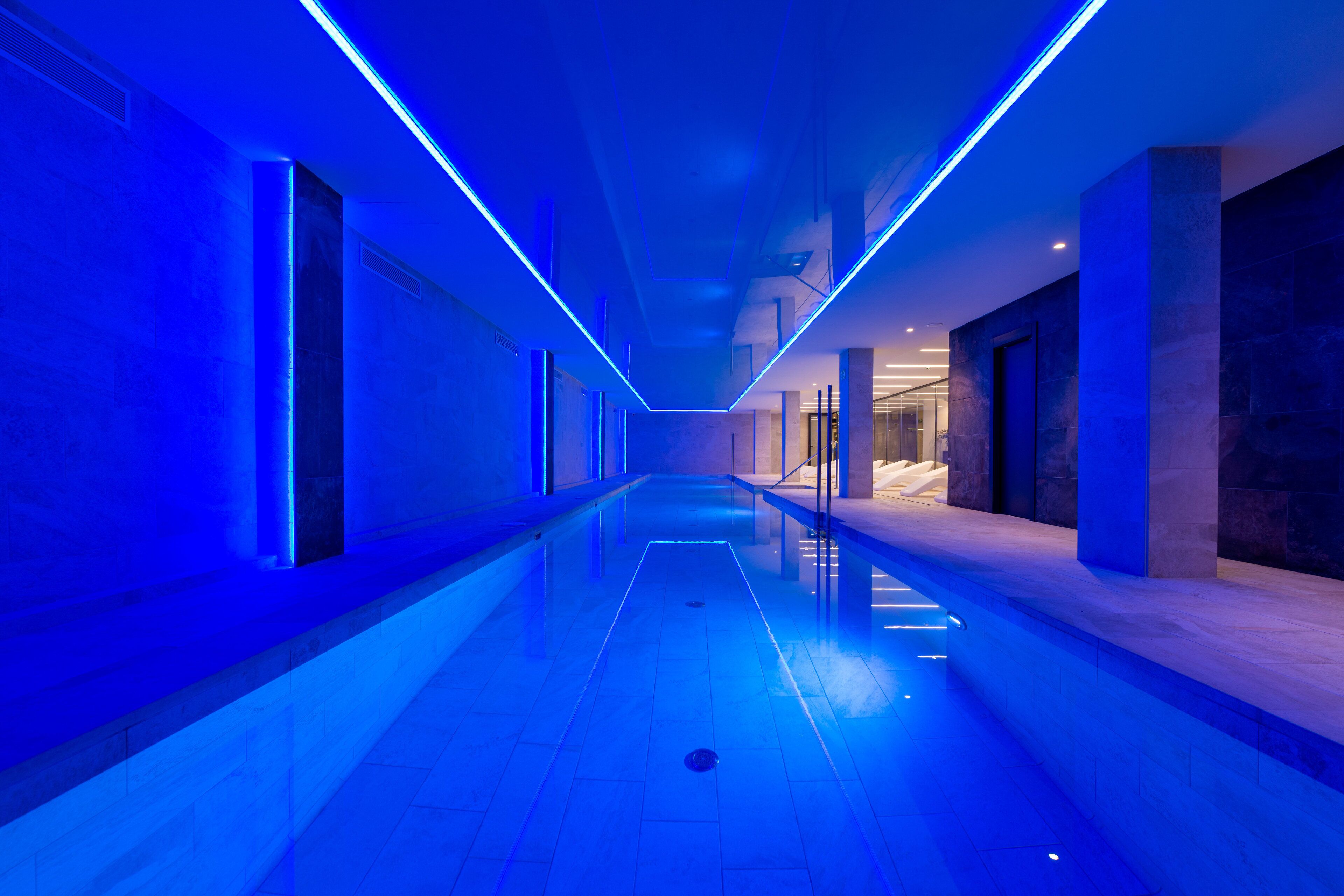 Un pasillo moderno con piso reflectante, iluminación azul y un ascensor de vidrio.