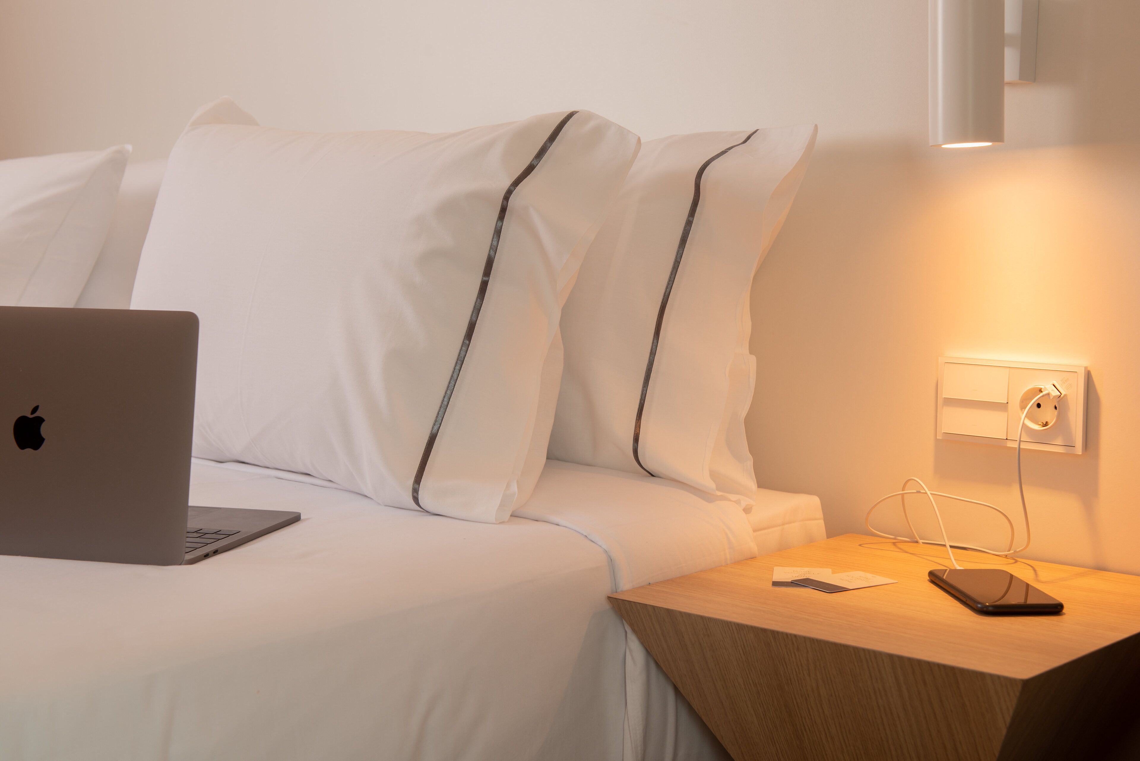 Un letto rifatto con lenzuola bianche e un laptop sopra. Un comodino con uno smartphone e un caricabatterie collegato.