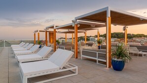Un área de lounge en la azotea con sillones blancos, pérgolas de madera y plantas en macetas.