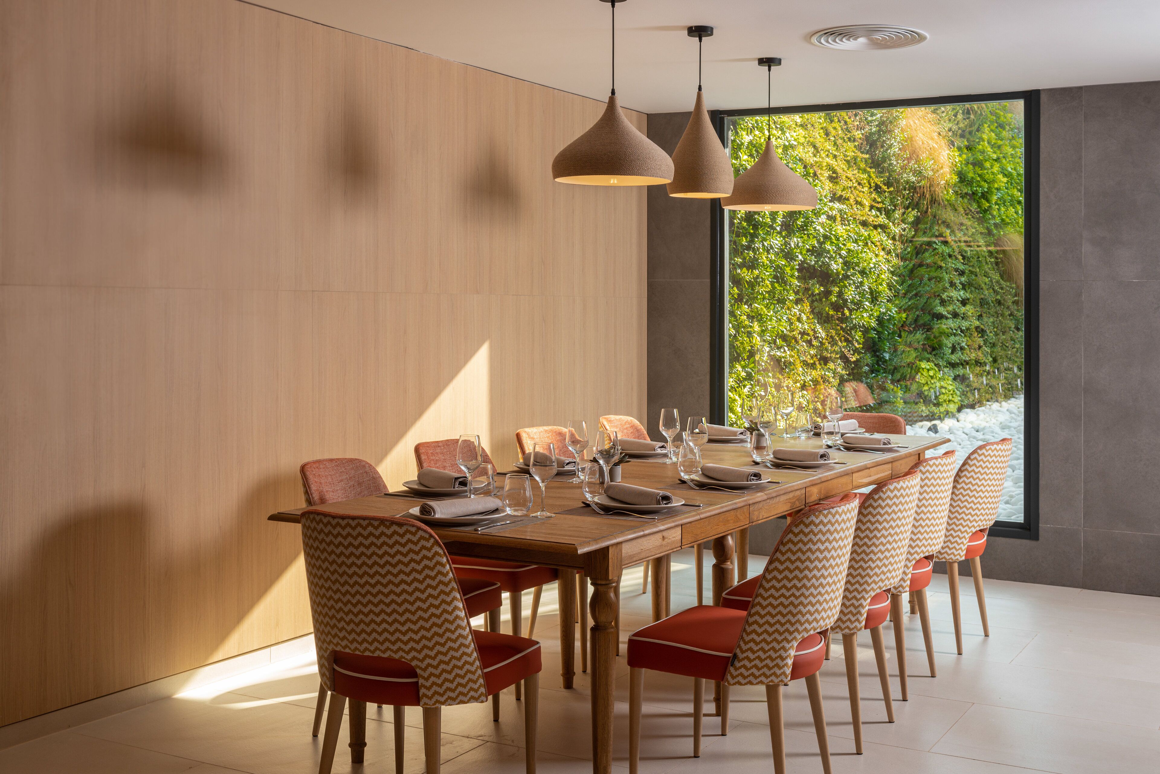 A modern dining area with a wooden table set for six, surrounded by patterned chairs, with pendant lighting and a view of greenery through large windows.