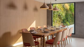 A modern dining area with a wooden table set for six, surrounded by patterned chairs, with pendant lighting and a view of greenery through large windows.