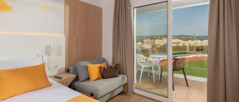 Habitación de hotel con cama, sofá, una mesita y vista a un campo deportivo a través de un ventanal.