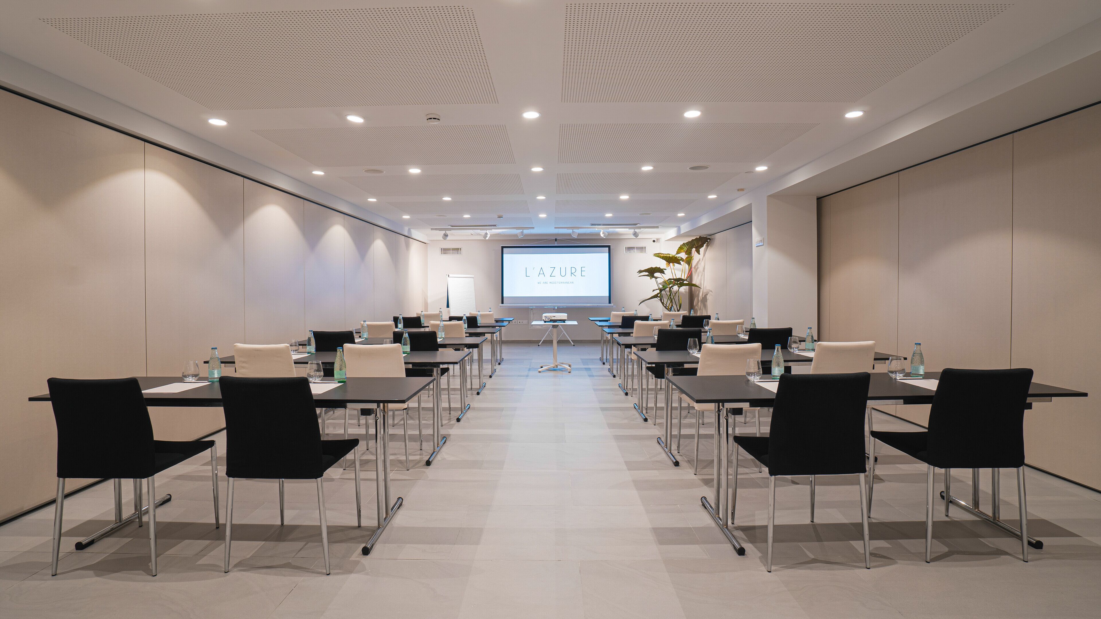 A conference room with a large screen displaying 'L'AZURE WE ARE MEDITERRANEAN'. Tables are arranged with chairs, and water bottles are on the tables.