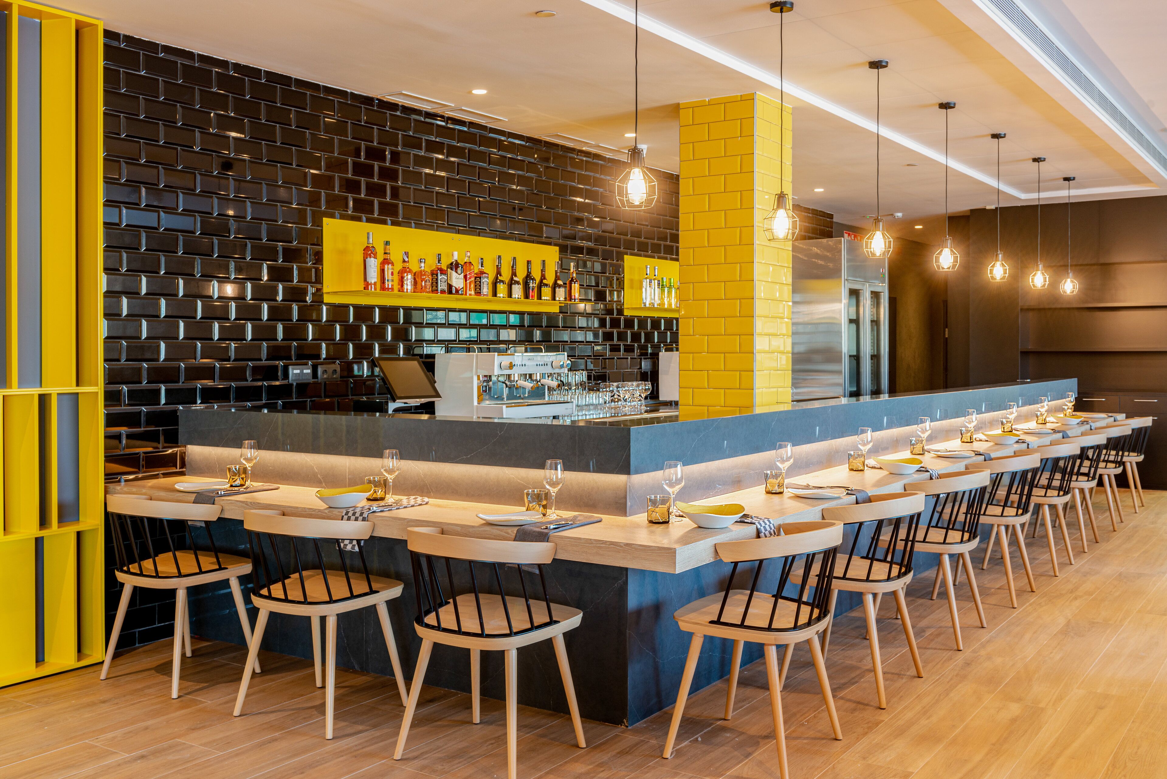A modern bar area with a long counter, multiple stools, and a brick wall backdrop.