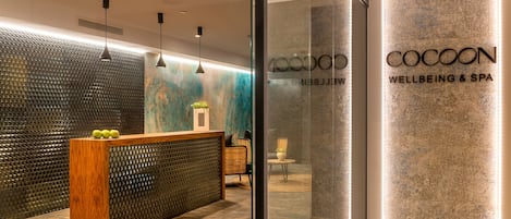 Un área de recepción con un mostrador de madera, una pared texturizada y un panel de vidrio con el nombre 'COCOON WELLBEING & SPA'.
