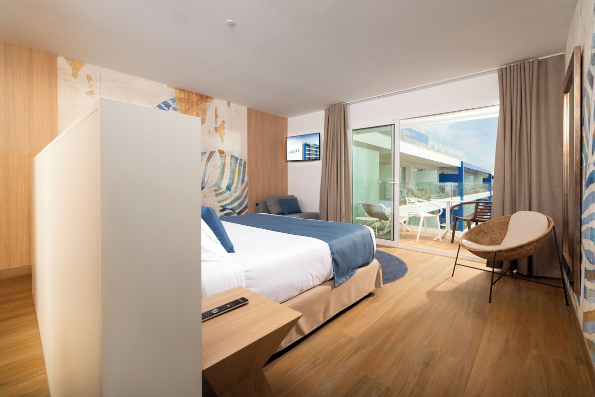 Una habitación de hotel moderna con cama, una mesita, una silla y un ventanal que da a un balcón con mobiliario exterior.