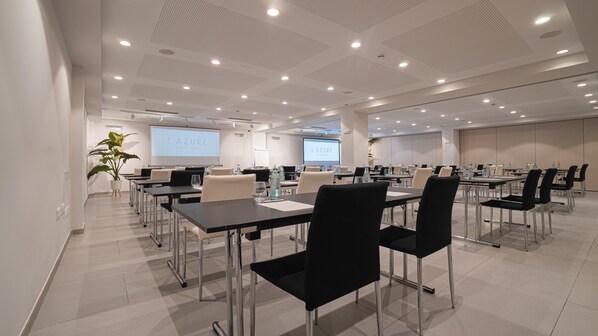 A conference room with multiple tables and chairs, a large screen displaying 'L'AZURE WE ARE MEDITERRANEAN', and a potted plant. - L'Azure Hotel (Lloret de Mar)