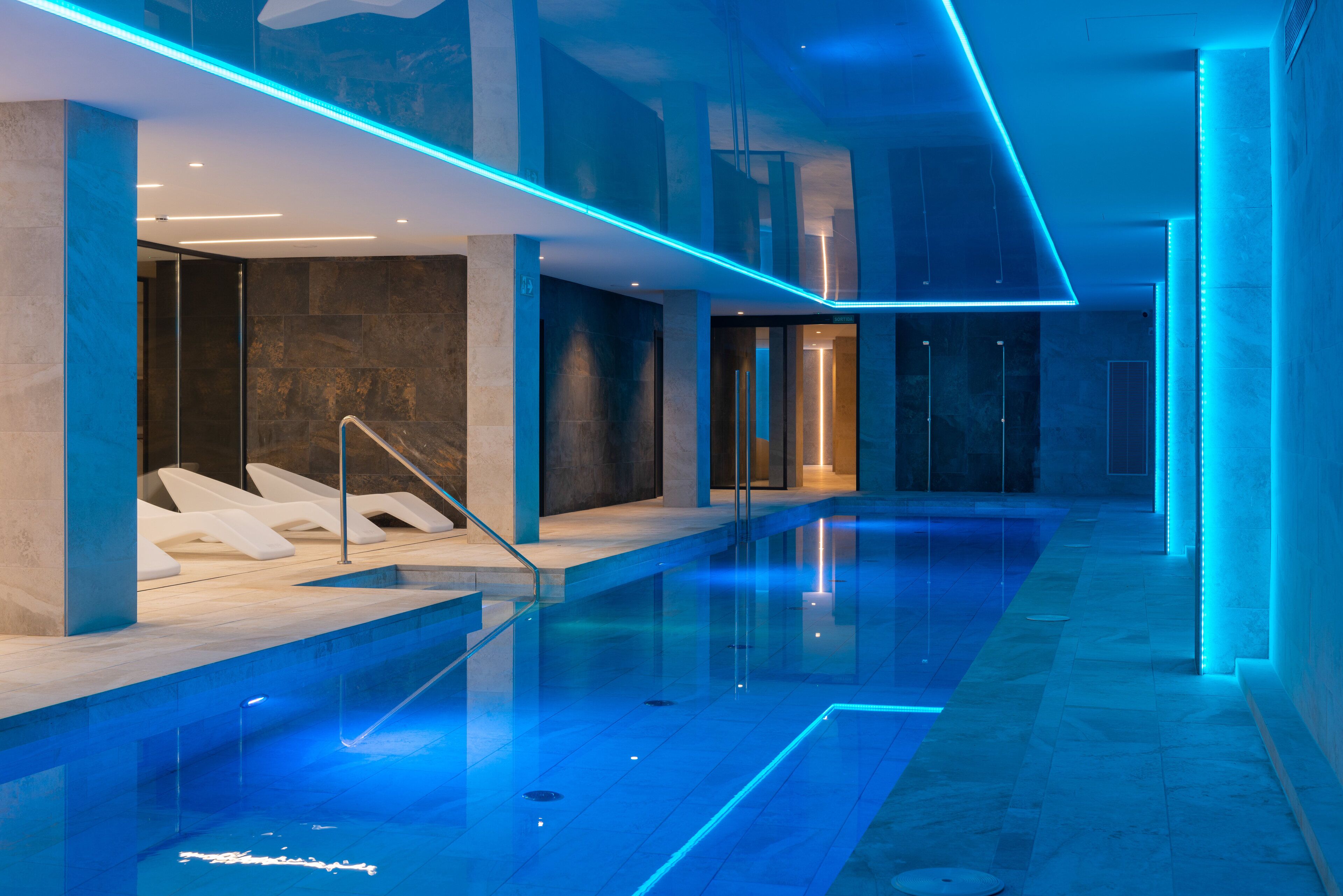 A modern indoor swimming pool with blue lighting, clear glass walls, and a minimalist design.