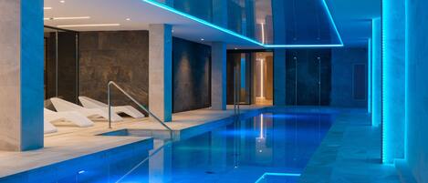 Una piscina interior moderna con iluminación azul, paredes de vidrio transparente y un diseño minimalista.