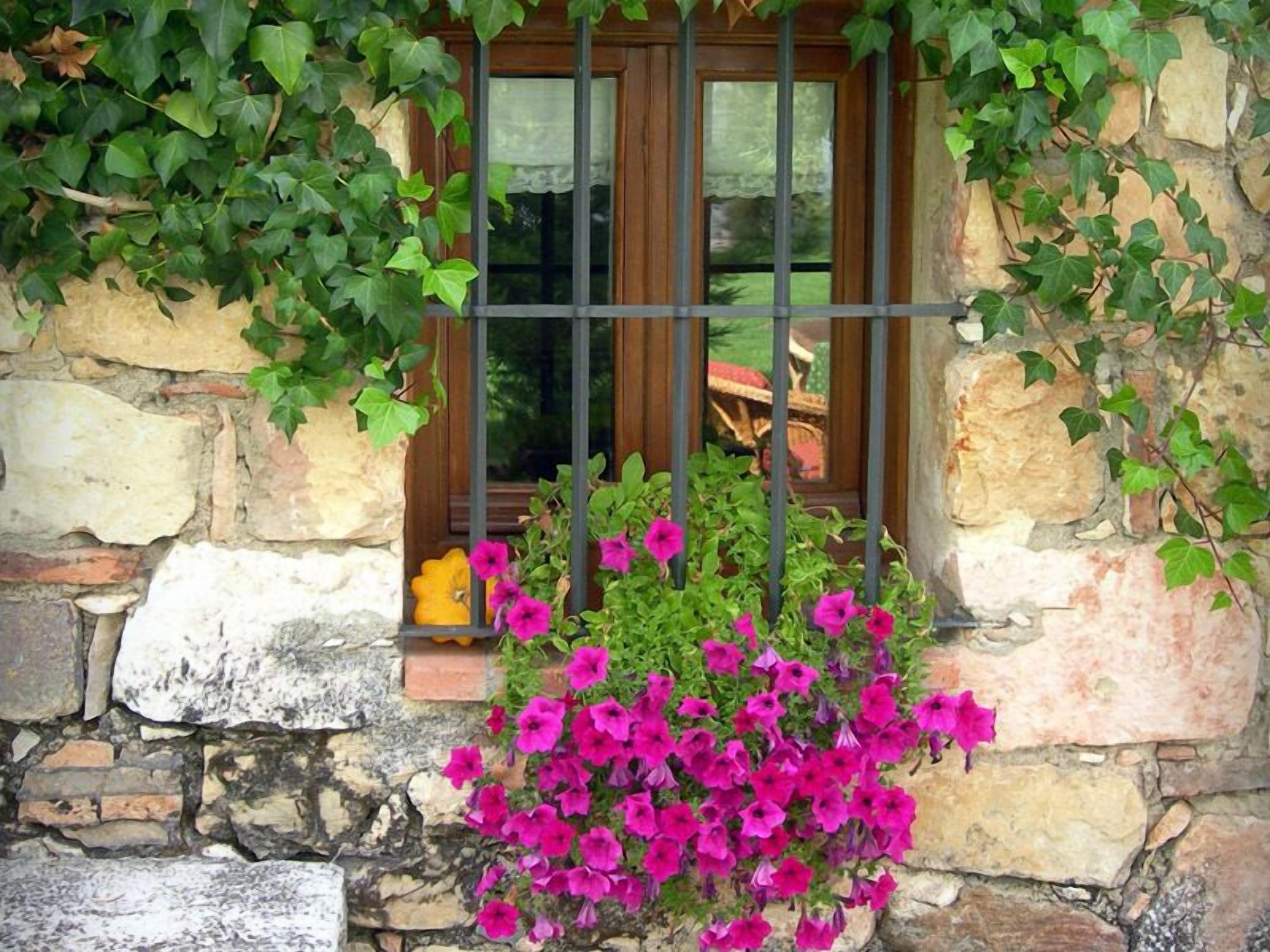 Un muro de piedra con una ventana que tiene barrotes de metal, una maceta amarilla y flores rosas.