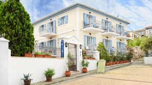 A two-story building with a white facade, blue shutters, and a balcony with a metal railing. There are potted plants along the front and a signboard visible.