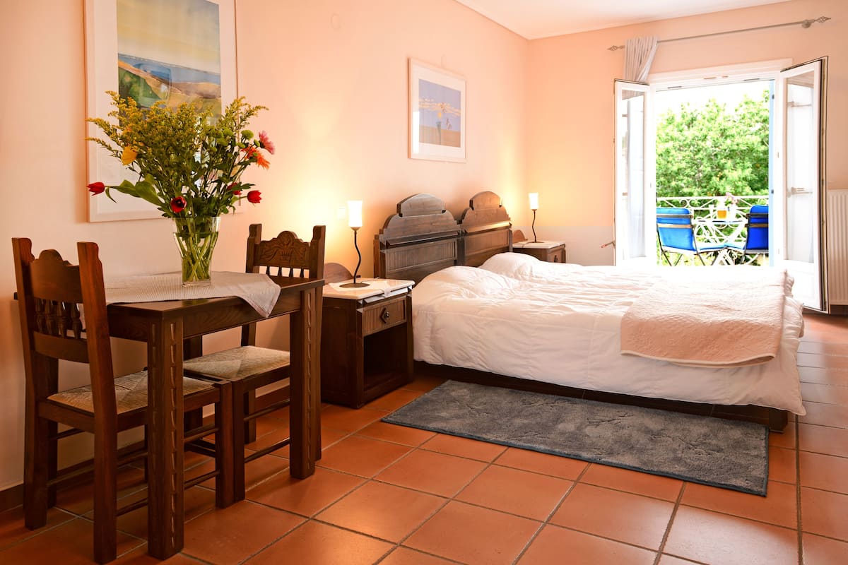 A bedroom with a bed, a desk with a chair, a small table, a vase with flowers, and a window with a view of the outside.
