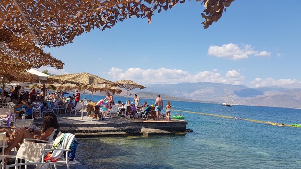 A beachside café with outdoor seating, a dock extending into the water, and a sailboat in the background.