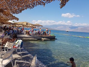 A beachside café with outdoor seating, a dock extending into the water, and a sailboat in the background. - Mira Mare Hotel (Delphi)