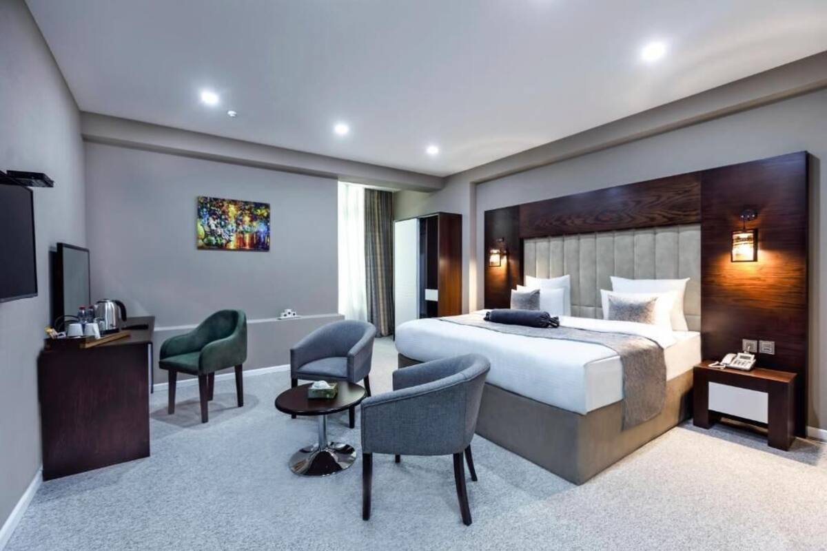 A modern hotel room with a large bed, two armchairs, a small table, a desk with a TV, and a painting on the wall.