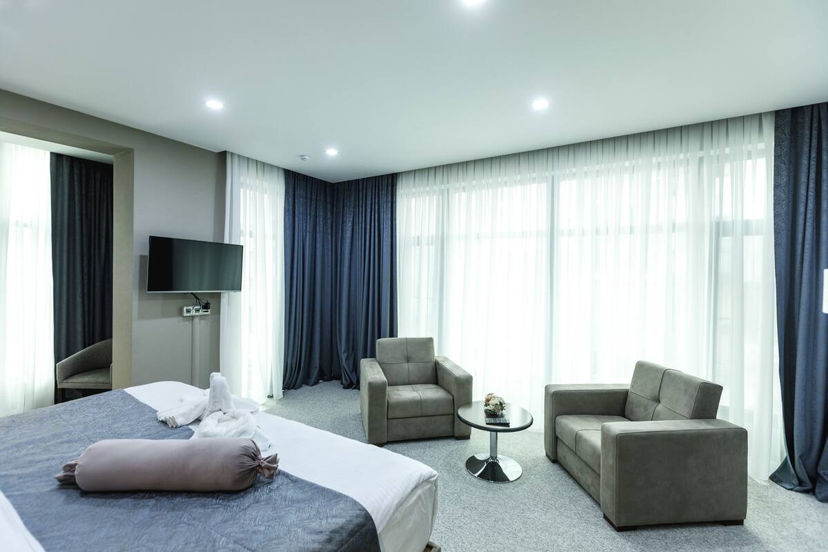 A modern hotel room with a large bed, two armchairs, a small round table, a flat-screen TV, and large windows with sheer curtains.