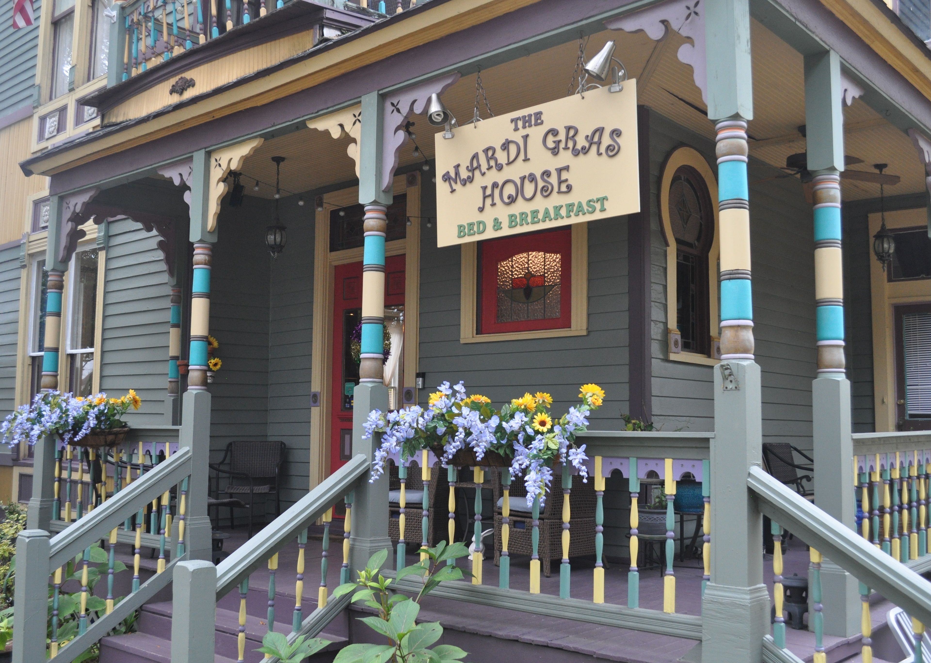 A historic building with a porch, a sign reading "The Mardis Gras House Bed & Breakfast," and a flower arrangement.