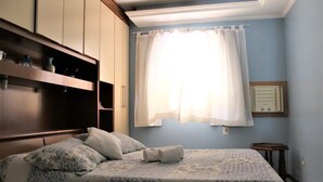 Un dormitorio con cama, armario, ventana con cortinas y una mesita.