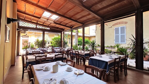 Un área de comedor al aire libre con techo, con varias mesas y sillas, rodeada de plantas y vistas a la calle.