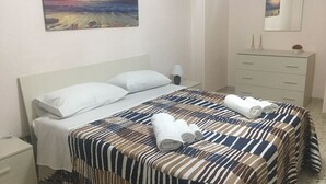 A neatly made bed with striped bedding, a nightstand with a lamp, a dresser, and a painting of a sunset over the ocean on the wall. - Casa Vacanze Da Angela (Sciacca)