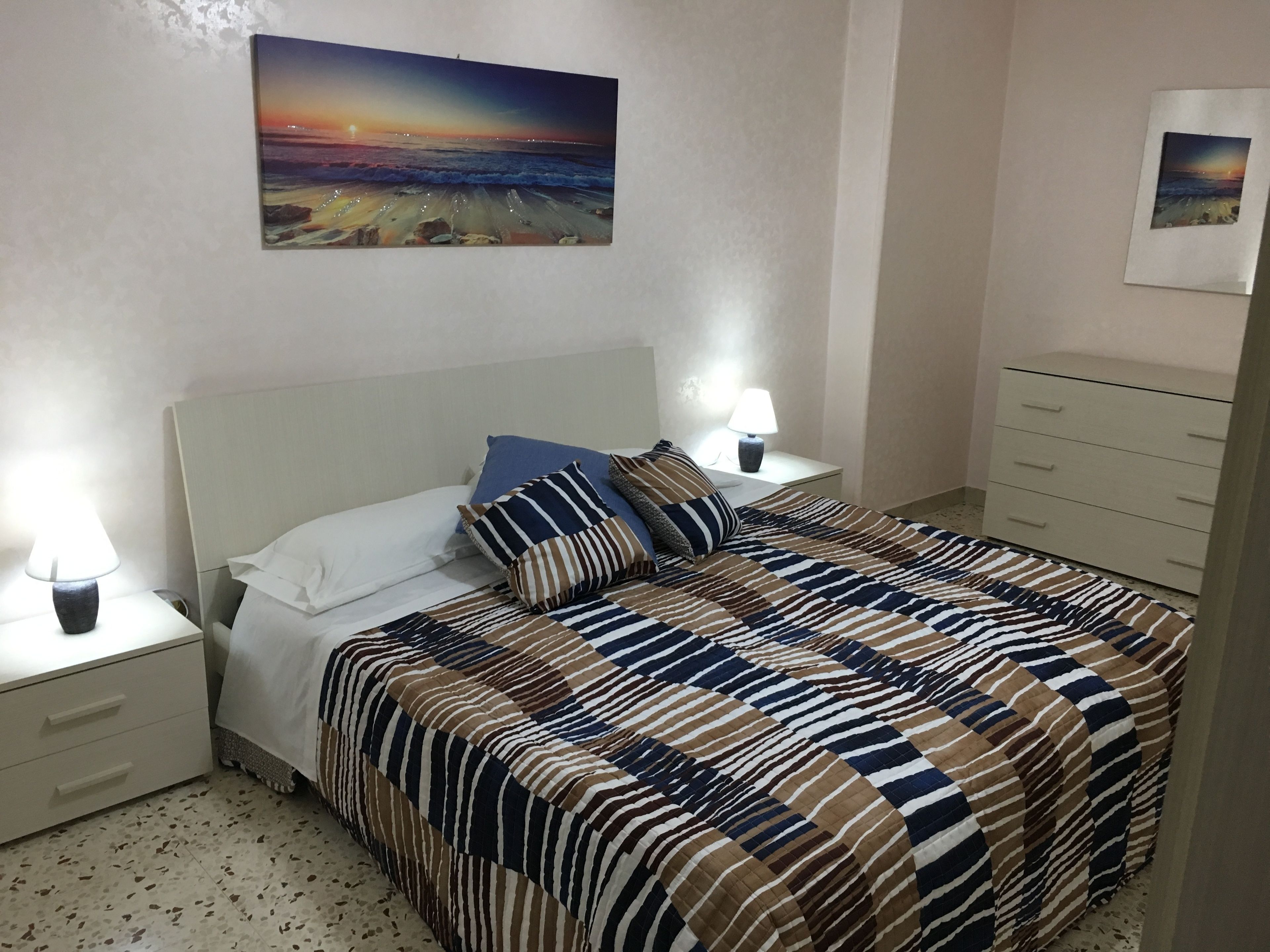Un letto rifatto con biancheria a righe, un comodino con una lampada e un quadro raffigurante un tramonto sulla parete.