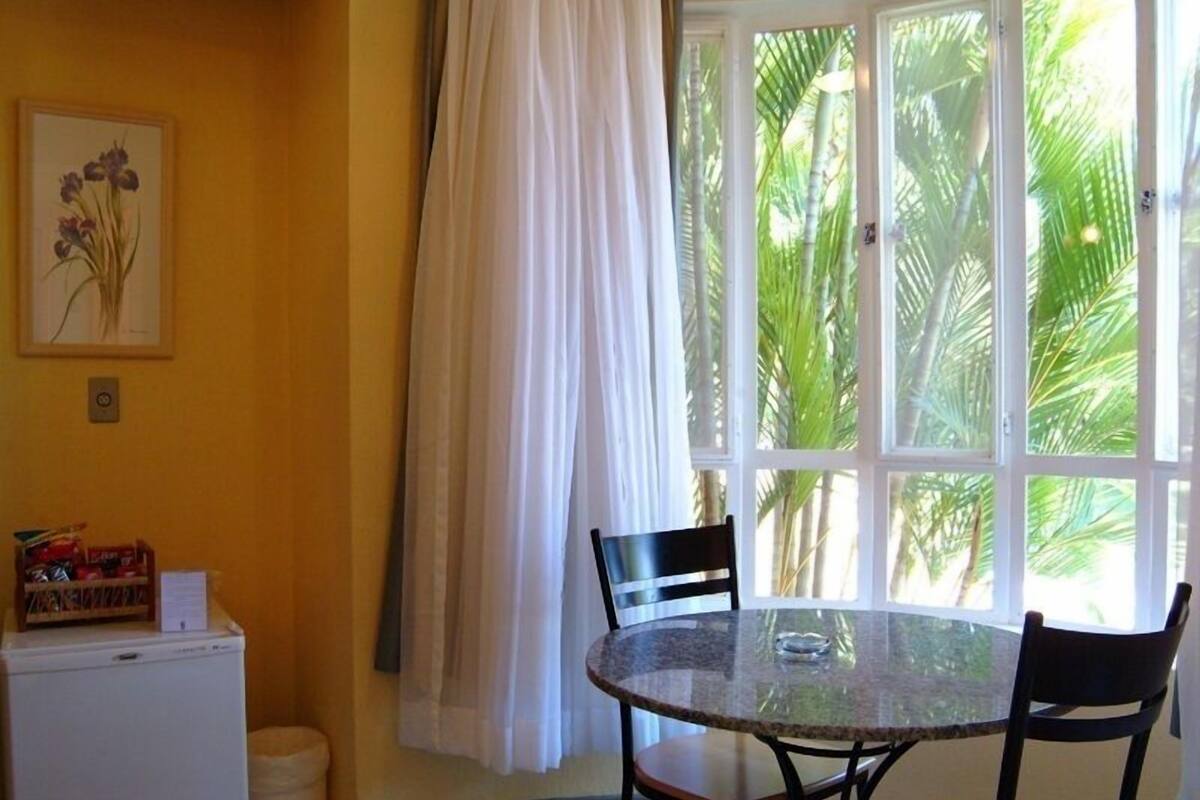 A small dining area with a round glass table and two chairs, a white refrigerator, a trash bin, and a window with a view of palm trees.