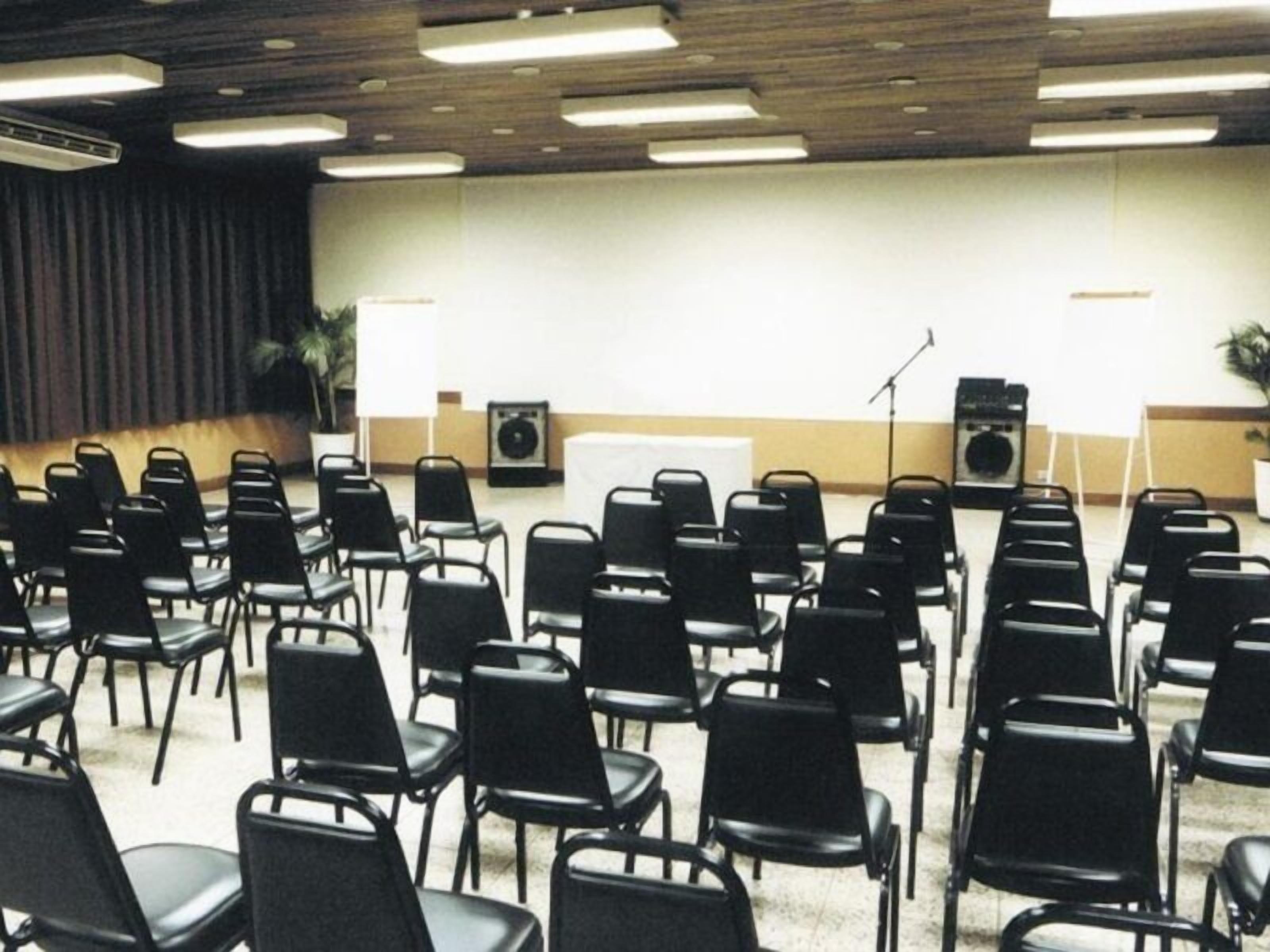 A conference room with rows of black chairs, a podium, a whiteboard, and two speakers.