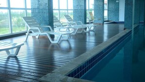 A swimming pool area with lounge chairs and a view of greenery outside. - Lindo Amplo 3 Quarto em Prédio Frente Mar (Cabo Frio)