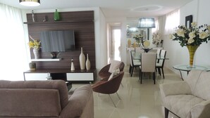 A modern living room with a flat-screen TV, a dining area with a glass table, and a kitchen in the background. - Lindo Amplo 3 Quarto em Prédio Frente Mar (Cabo Frio)