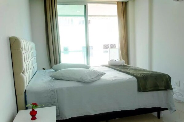 A hotel room with a bed, a white nightstand, a red vase with flowers, and a view of the outside through a window.