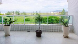 A balcony with a view of a green landscape, two potted plants, and a tiled floor. - Lindo Amplo 3 Quarto em Prédio Frente Mar (Cabo Frio)