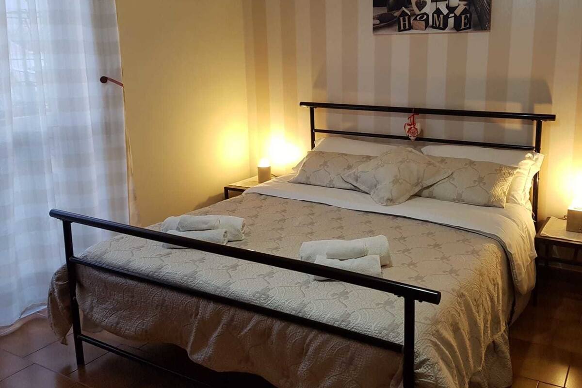 Ein ordentlich bezogenes Bett mit Metallgestell, zwei Nachttische mit Lampen und ein Wandbild.