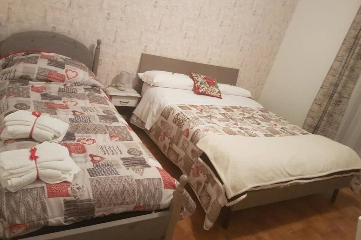 Ein Schlafzimmer mit zwei Betten, jeweils mit gemusterten Bettdecken und Kissen. Dazwischen steht ein Nachttisch mit einer Lampe.
