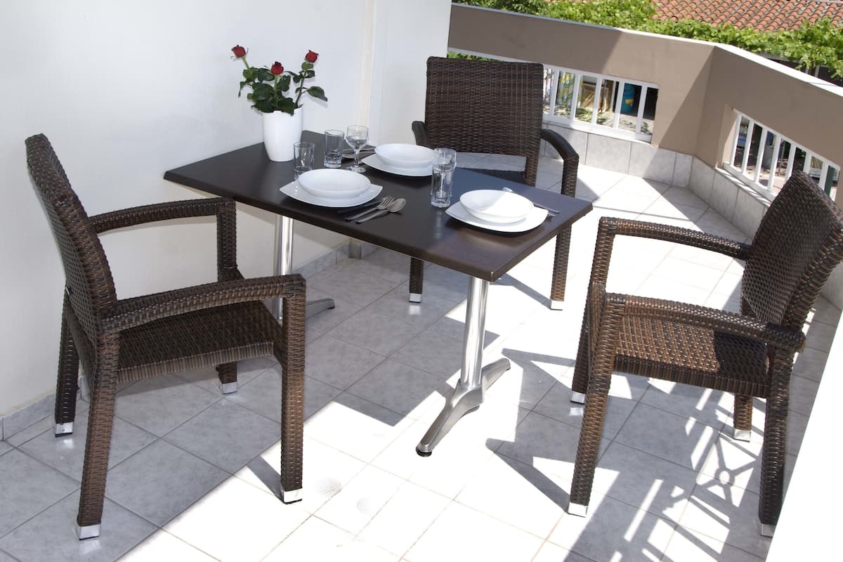 A dining table set with plates, glasses, and cutlery on a balcony with wicker chairs and a potted plant.