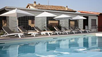 Ein Poolbereich mit Liegestühlen und Sonnenschirmen, ein klares Schwimmbecken und im Hintergrund ein Gebäude mit einem Ziegeldach.