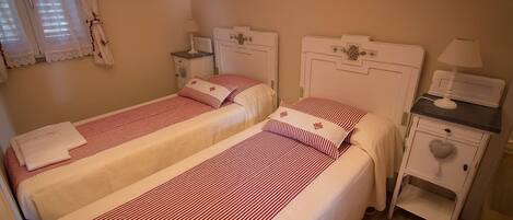 Habitación con dos camas, cada una con cabecero blanco y ropa de cama a rayas rojas. Hay una mesita de noche con una lámpara y un estante pequeño.