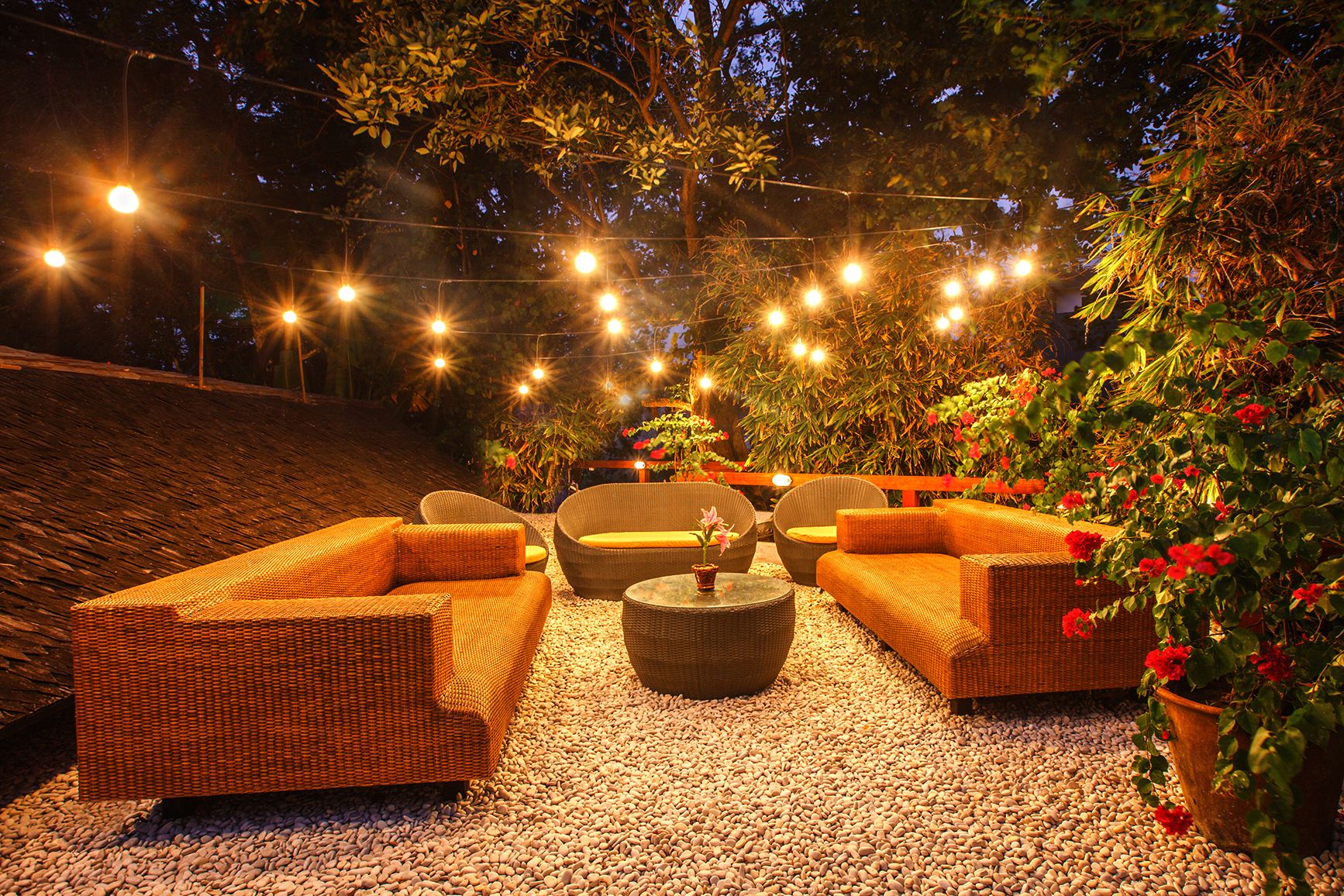 Un área de estar al aire libre con mobiliario de mimbre, luces colgantes y plantas en macetas.