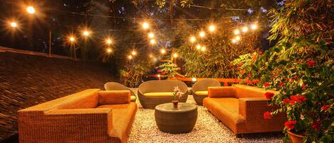 Un área de estar al aire libre con mobiliario de mimbre, luces colgantes y plantas en macetas.