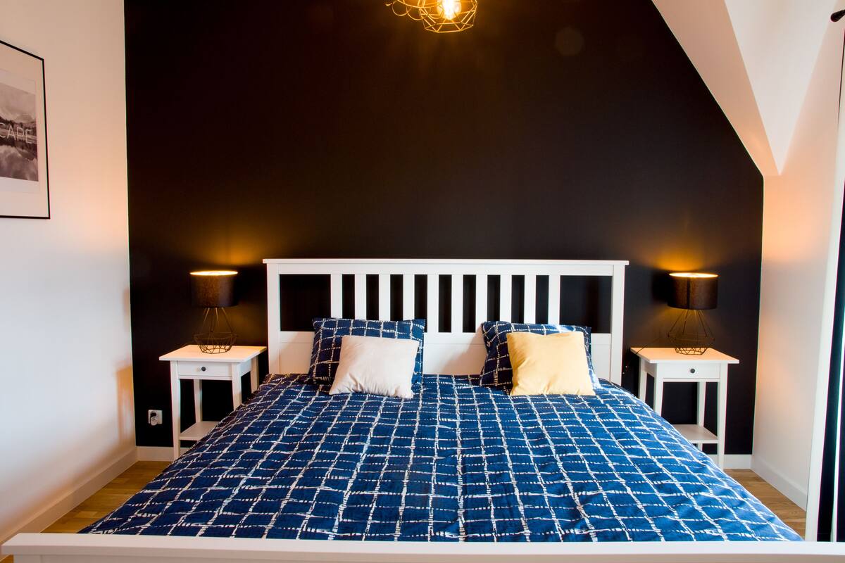 A bedroom with a white headboard, a blue patterned bedspread, two bedside tables with lamps, and a framed picture on the wall.