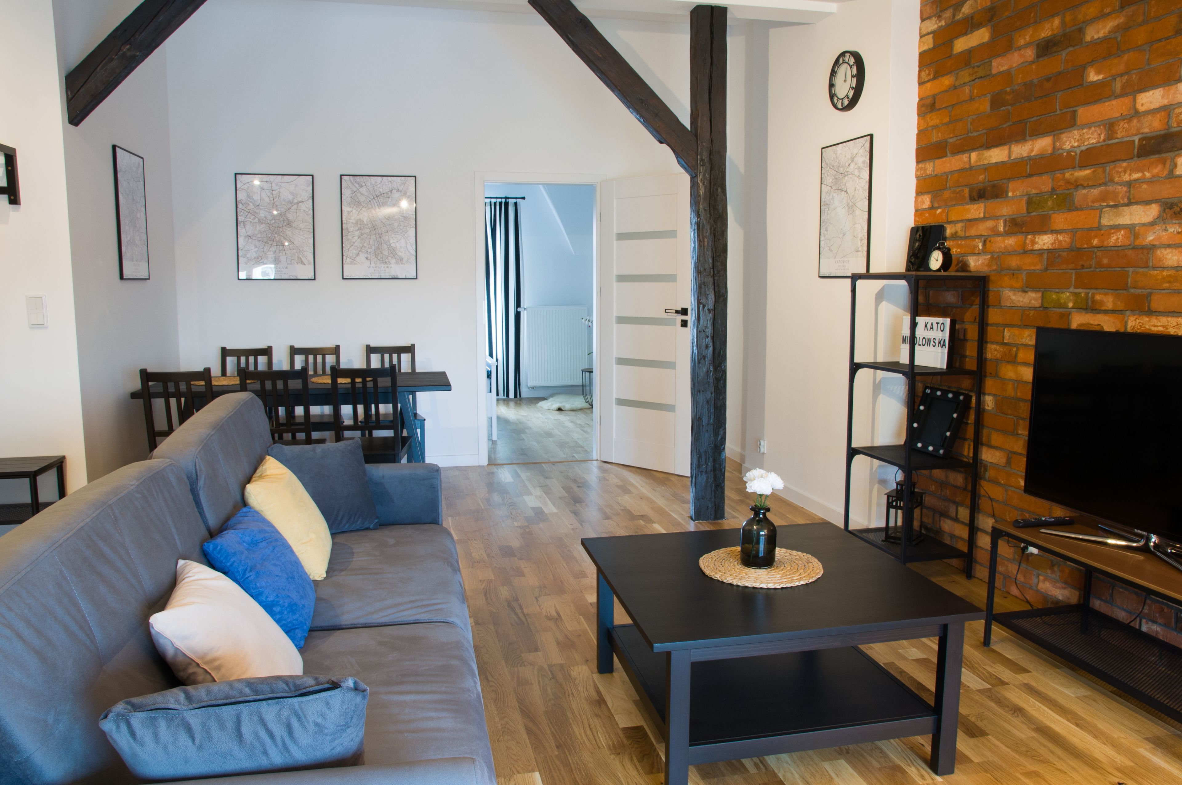 A living room with a sofa, coffee table, and TV. There is a brick fireplace and a dining area in the background.
