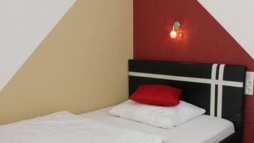 Una cama con estructura negra y ropa de cama blanca, un almohadón rojo y una lámpara de pared.