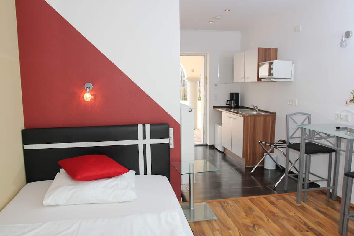A modern hotel room with a bed, a red pillow, a glass table, and a kitchenette in the background.