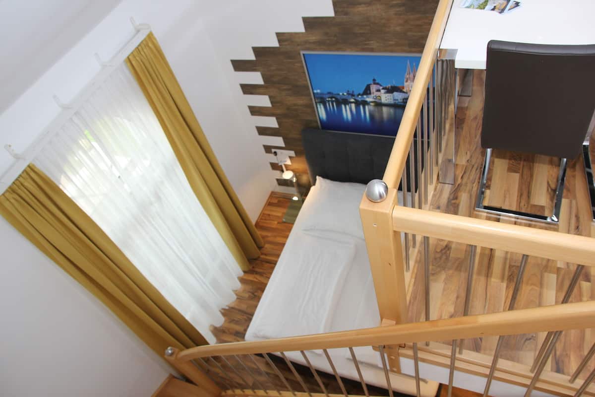 A staircase with a wooden handrail, a white mattress on the floor, a black chair, and a wall-mounted TV displaying a cityscape.