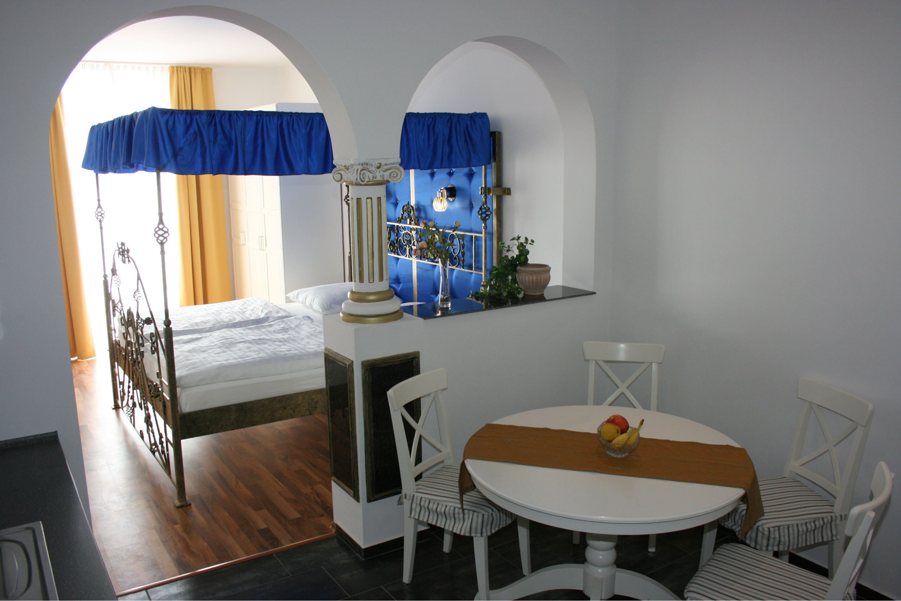 A bedroom with a bed, a small table with a fruit bowl, and a window with curtains.