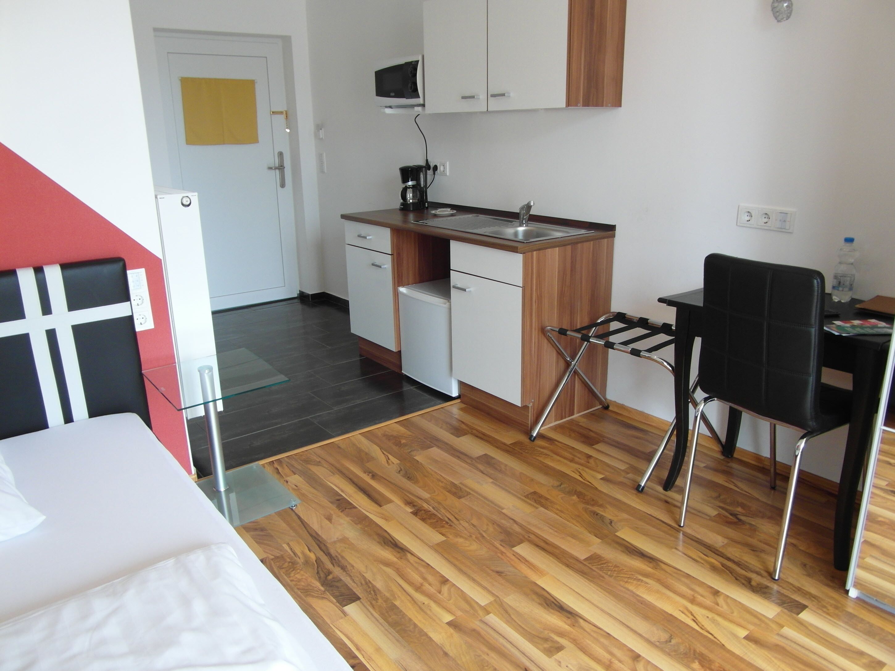 Una cocina compacta equipada con microondas, cafetera y lavabo. Se observa un armario de madera y una silla negra. En primer plano, se ve una cama.