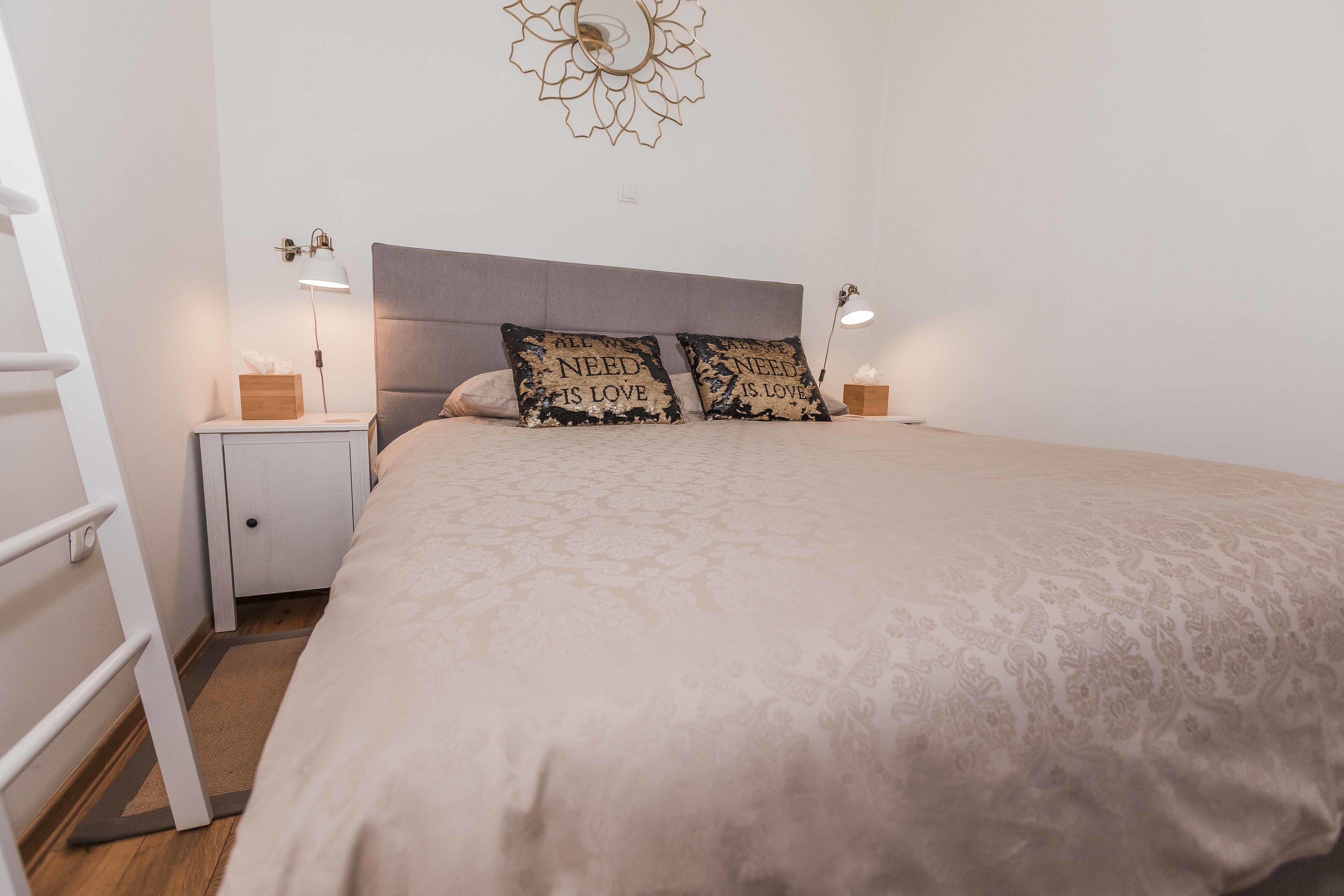 Cama bem arrumada com colcha estampada, duas luminárias de cabeceira, mesa de apoio com caixa de lenços e um objeto decorativo acima da cama.