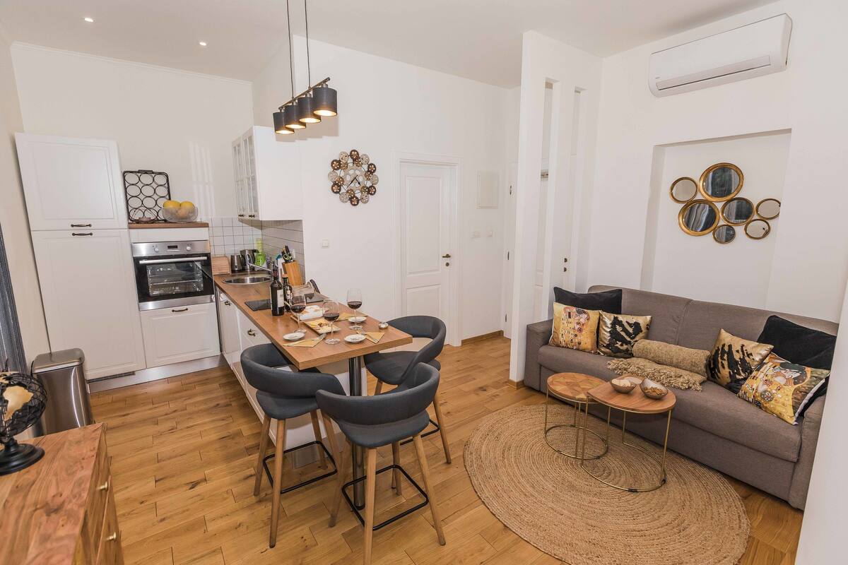 A modern kitchen with white cabinets, a dining area with a wooden table and chairs, and a living room with a sofa and decorative mirrors.