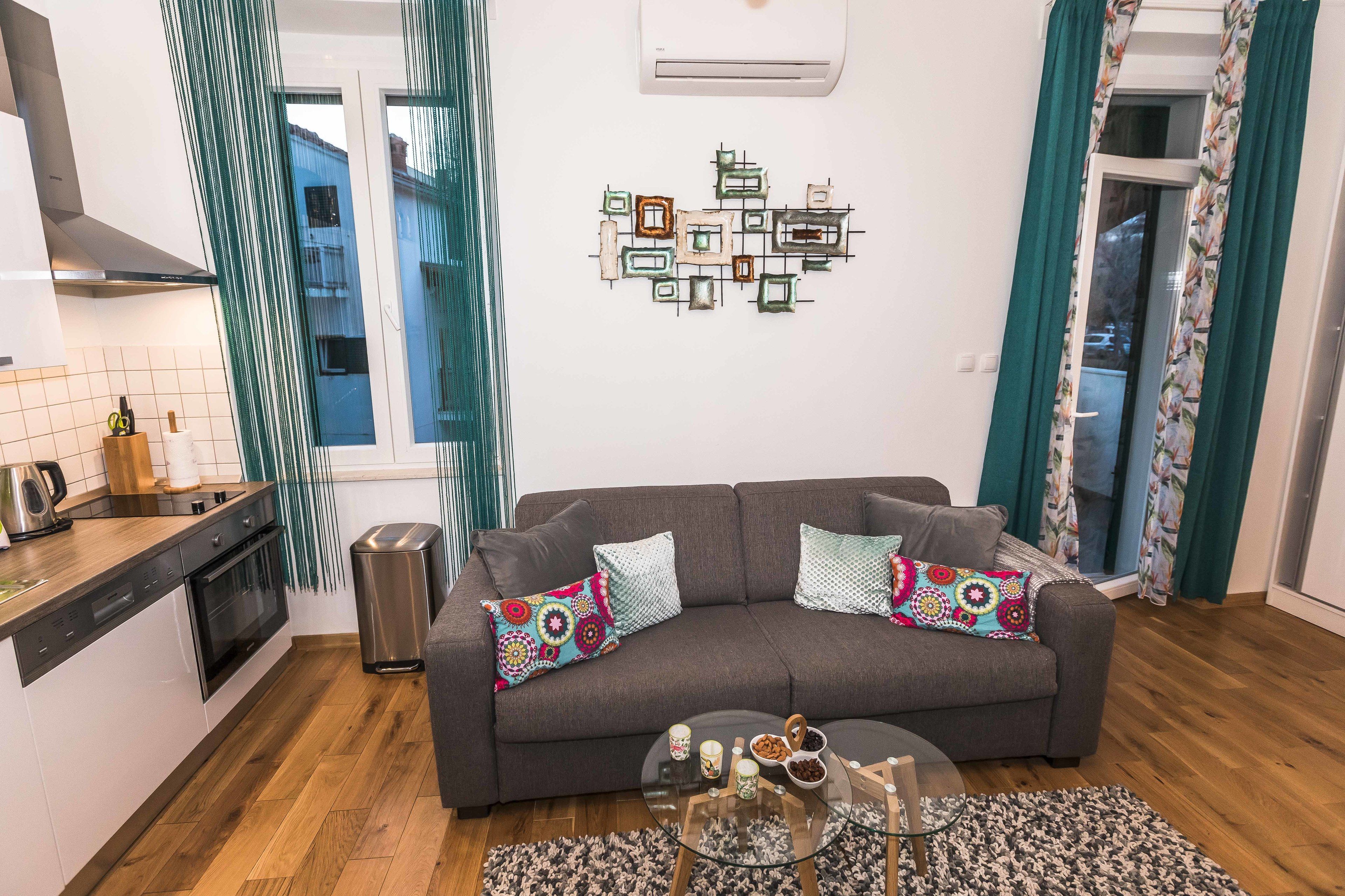 A modern living room with a grey sofa, a coffee table, and a kitchen area with a dishwasher and oven.