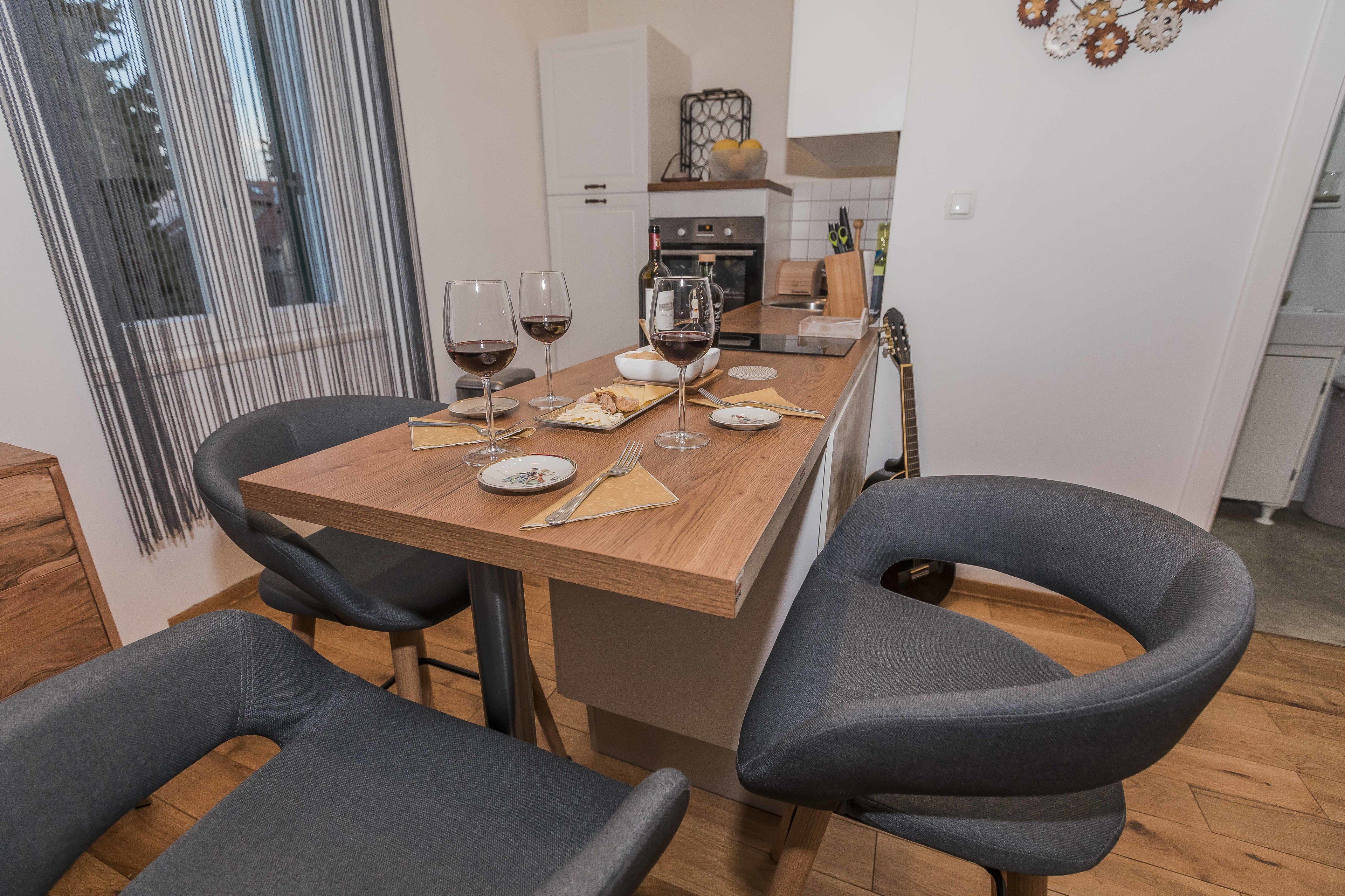A dining area with a wooden table, grey upholstered chairs, and a kitchen in the background.